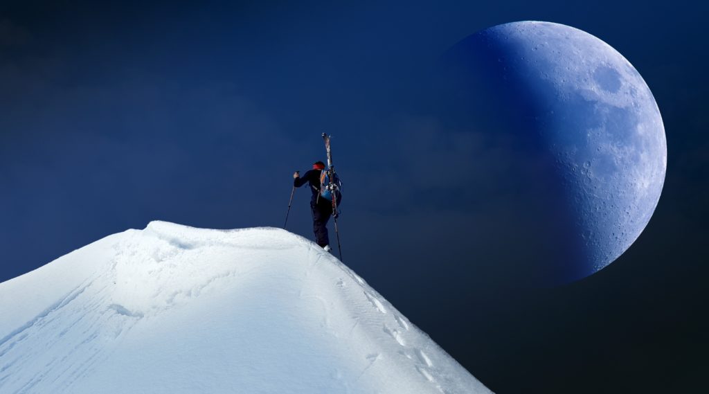 Skier summiting mountain at night with moon in backdrop